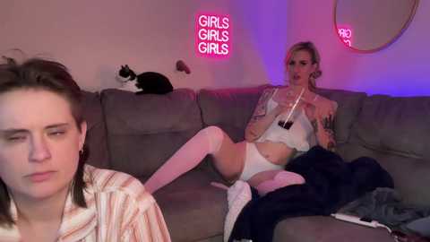 Video of two transgender women in a cozy living room; one lounges on a gray couch, wearing white lingerie and thigh-high socks, while the other, in a striped shirt, sits nearby. A black-and-white cat sleeps on a cushion.