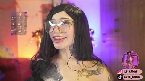 Video of a smiling woman with long black hair, wearing glasses and a tattoo on her chest, standing in a dimly-lit room with a purple and yellow background.