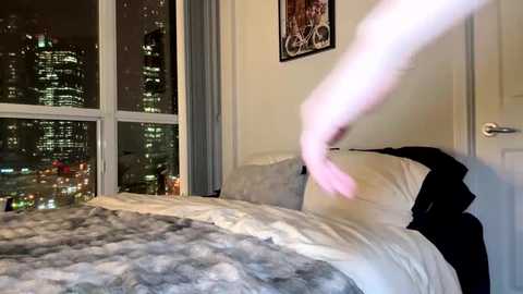 Video of a modern bedroom with a white bed, gray and white fur blanket, and cityscape view through large windows at night. A ghostly, floating arm in white extends from the bed, adding a supernatural element.