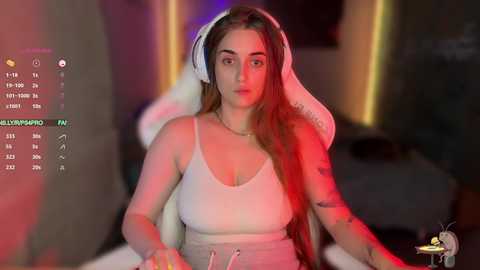 Video of a young, light-skinned woman with long, straight brown hair in a white tank top, wearing headphones. She sits in a gaming chair, surrounded by dim lighting with neon accents.