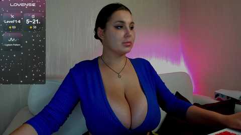 Video of a curvy woman with large breasts in a plunging blue top, sitting on a white couch. She has fair skin and dark hair. Background shows a TV displaying a Love Island UK episode.