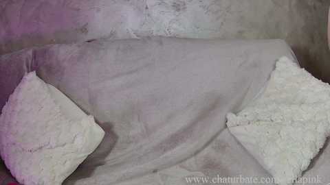 Video of a gray, textured fabric draped over a surface, featuring two white, feather-like pillows. The background is a soft, muted palette with subtle purple and gray hues, creating a serene, minimalist atmosphere.