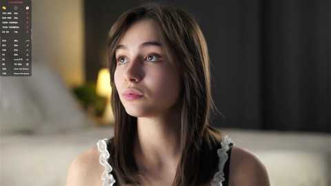 Media: Video of a young Caucasian woman with fair skin, long brown hair, and blue eyes, wearing a white and black ruffled top, looking contemplative in a dimly-lit, modern room.