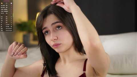 Video of a young woman with fair skin, straight black hair, and a slim build, wearing a red bra, standing in a modern bedroom with white bedding and a green potted plant.