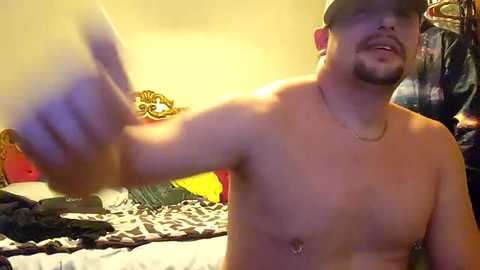 Video of a shirtless man with a medium build and light skin tone, giving a thumbs-up. He has a trimmed beard and wears a black baseball cap. The background shows a messy bed with colorful pillows and a yellow wall.