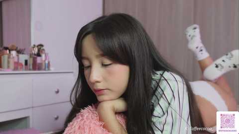 Video of a young Asian girl with long black hair, lying on her stomach in a room, wearing a striped shirt and white socks, with a fluffy pink pillow. Makeup and skincare products on a dresser in the background.