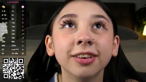 Video of a young woman with fair skin, dark hair, and a septum piercing, smiling. Background shows a blurred, dark room with a bed. QR code in lower left corner.