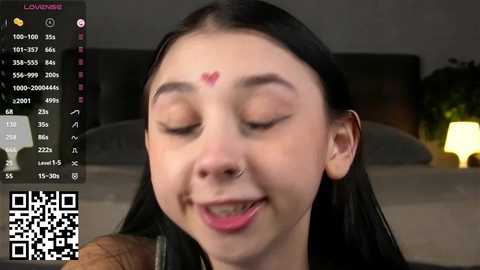 A video of a young woman with long black hair, closed eyes, and a heart-shaped mark on her forehead, smiling playfully. She's indoors, with a blurred background featuring a bed and a potted plant.