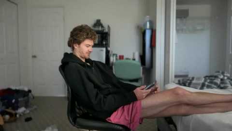 Video of a young man with curly hair, wearing a black hoodie and pink shorts, sitting on a black chair in a messy bedroom, focused on his phone.