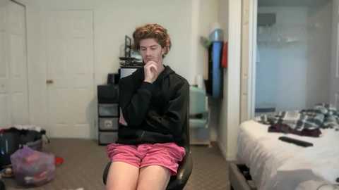 Video of a young person with curly brown hair, wearing a black hoodie and pink shorts, sitting pensively on a chair in a messy bedroom with scattered clothes, a TV, and a door.