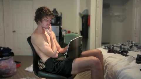 A video of a shirtless, muscular young man with curly hair, wearing black shorts, sitting on a chair in a cluttered, dimly-lit bedroom, typing on a laptop.