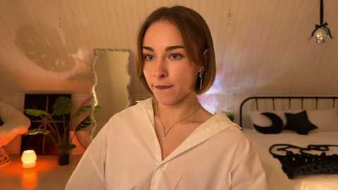 A video of a young woman with short brown hair, wearing a white robe, standing in a dimly-lit, cozy bedroom with a bed, potted plants, and a candle.