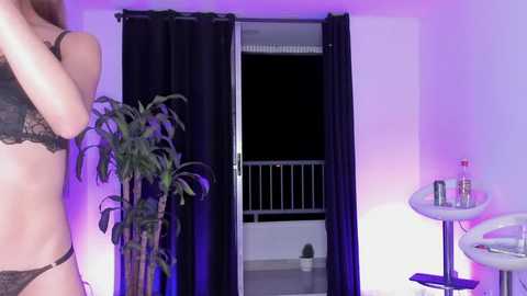 Video of a woman in black lace lingerie, standing near a partially open balcony with dark curtains. A potted cactus and a small table with bottles are visible in the background.