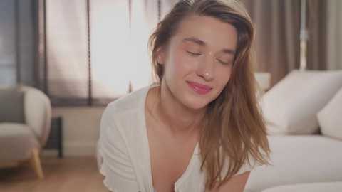 Media: Video of a smiling young woman with long, light brown hair, wearing a white blouse, in a modern, softly lit living room with white furniture and beige curtains.