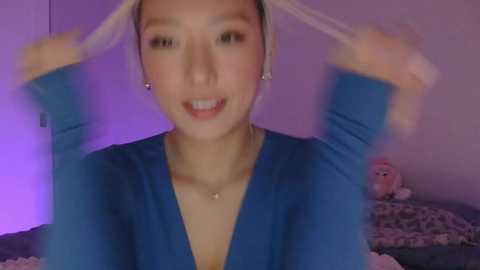 Video of a young Asian woman with light skin, wearing a blue V-neck top, adjusting her hair, smiling, in a dimly lit bedroom with a teddy bear.