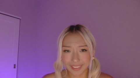 Video of a smiling Asian woman with long, blonde hair in pigtails, wearing minimal makeup, against a purple-tinted wall.