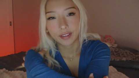 A video of an Asian woman with long, platinum blonde hair, wearing a blue velvet dress, sitting on a bed with plush pillows. The background has a red light and a stuffed animal.