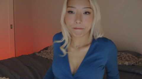Video of a blonde, light-skinned woman with a V-neck blue top, sitting on a bed with patterned pillows. Background features a closed door and soft, warm lighting.