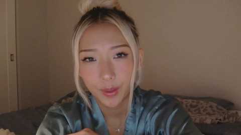 Video of an Asian woman with light skin and platinum blonde hair tied in a bun, wearing a teal satin robe. She's puckering her lips, in a dimly lit bedroom with beige walls.