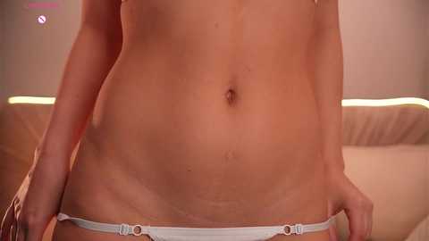 Video of a woman's torso, from mid-thigh to mid-chest, showing a white thong with metallic rings, fair skin with visible stretch marks, and a soft-focus background of a beige sofa.