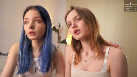 Video of two young women with pale skin and blue hair (left) and brown hair (right). Both wear casual, sleeveless tops; background shows a blurred indoor setting with warm lighting.