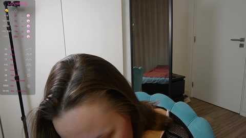 Video of a young woman with straight, shoulder-length brown hair, wearing a black top, partially obscured by a blue headband. Background includes a wardrobe, a bed with a red blanket, and a wooden floor.