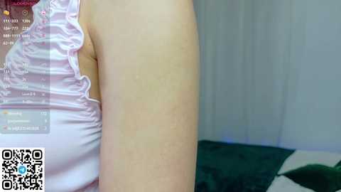 A close-up video of a person's bare shoulder and upper arm, showing fair skin and a pastel pink sleeveless top with ruffled details. The background features a bed with a dark green blanket and white sheets.