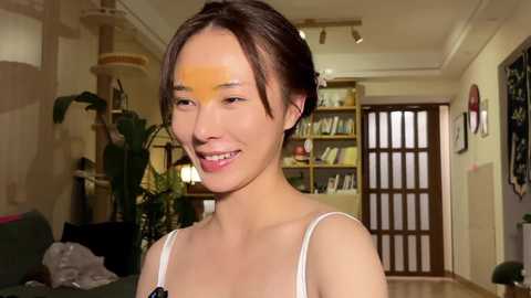 Video of an East Asian woman with light skin, dark hair in a bun, smiling, wearing a white spaghetti strap top. Background shows a modern living room with a bookshelf, plants, and a wooden door.