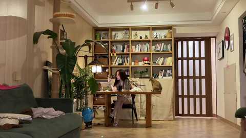 Video of a cozy, cluttered living room with a woman at a wooden desk, surrounded by bookshelves, a green couch, and a large plant.