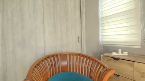 Video of a modern bedroom featuring a wicker chair with a turquoise cushion, a white wooden wardrobe, a light wooden dresser, and a window with white blinds, all bathed in natural light.