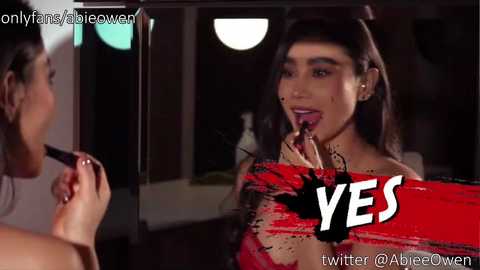 Media: Video: A woman with long dark hair, makeup, and red lipstick, wearing a red top, is applying makeup. Background: Dimly lit room with a mirror and circular lights. Text overlay: \"OnlyFans.com/AbieOwen\" and \"YES\".