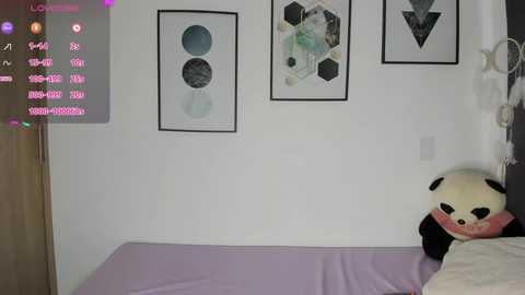 Video of a minimalist room with white walls, a bed covered in light lavender sheets, three abstract geometric art pieces, and a large, plush panda toy.