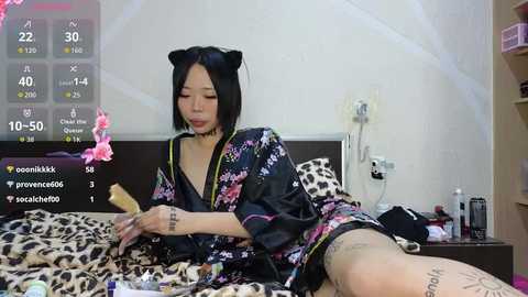 Video of an Asian woman with black cat ears and a floral kimono on a leopard-print bed, playing with makeup brushes in a messy bedroom.