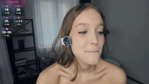 Video of a young, light-skinned woman with shoulder-length brown hair, wearing a gray scrunchie, and makeup brushes on her face, in a dimly lit room with a shelf in the background.