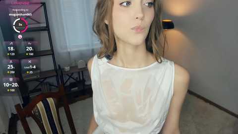 Video of a young woman with light skin and shoulder-length, wavy brown hair, wearing a sheer white top, making a pouty face, in a room with a black bookshelf, lamp, and striped chair.