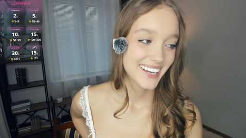 A young woman with long brown hair, wearing a white lace top, smiles while playing a video game in a modern living room with a TV and a bookshelf.