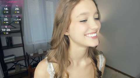 Video of a young, fair-skinned, smiling Caucasian woman with light brown hair, wearing a white lace top, indoors.