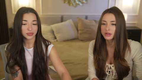 Video of two young Asian women with long black hair, wearing casual outfits, sitting in a modern bedroom with beige decor, smiling at each other.