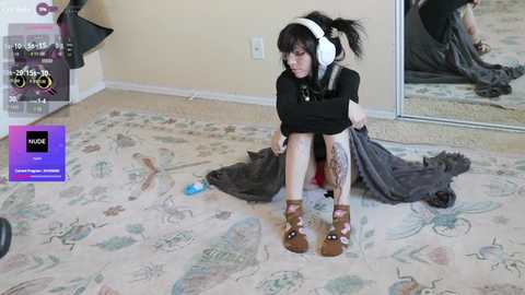 A video of a young woman with dark hair, wearing headphones, sitting on a carpeted floor, wearing a black shirt and gray skirt, surrounded by scattered clothes and a purple box.