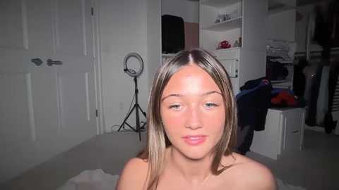 Video of a young, fair-skinned woman with long, straight brown hair, wearing a pink necklace, in a modern, white-walled bedroom with a studio light, clothes, and a mirror on the left.