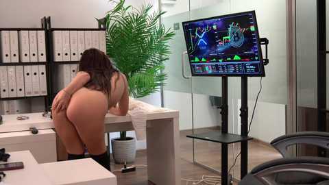 Video of a nude woman with long dark hair bent over a desk, facing a computer screen displaying a colorful game. Modern office setting with white furniture, green plants, and a black monitor.