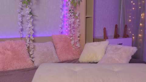 A video of a cozy bedroom with a plush pink and white faux fur sofa, adorned with white flower garlands and purple fairy lights, alongside a white dresser with wooden dildos.