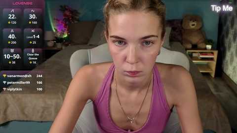 Video of a young woman with blonde hair, wearing a pink tank top, sitting on a bed in a dimly lit room, with a \"Tip Me\" screen and social media notifications visible.