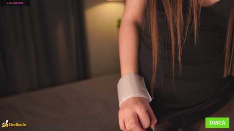 A close-up video of a woman with long brown hair, wearing a black sleeveless top and a white brace on her right arm, seated in a dimly lit room with dark curtains.