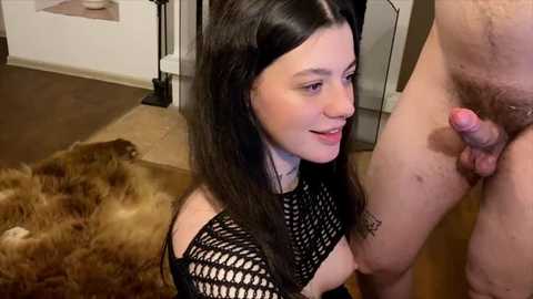 Video of a slender, fair-skinned woman with long black hair, wearing a black fishnet top, kneeling and smiling at a standing, naked man with a circumcised, erect penis. Background includes a beige couch, brown carpet, and a white wall.