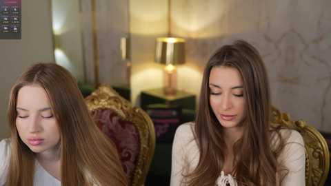 Video of two young women with long brown hair, one with closed eyes, sitting on a plush, gold-leafed sofa in a warmly lit room.