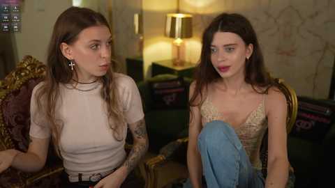 Video of two young women with long hair, one with a tattooed arm, sitting on a plush, ornate sofa in a dimly lit room with a vintage lamp and floral wallpaper.