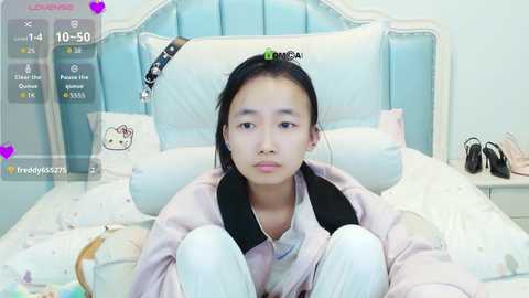 Media: A video of a young Asian girl with straight black hair, wearing a pink hoodie, sitting on a white bed with a light blue headboard, surrounded by plush toys, in a cozy bedroom.
