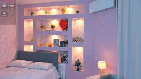 Video of a modern bedroom with a lavender wall featuring built-in shelves lit by soft lights, displaying green plants, books, and a red heart ornament. The bed has white linens and a gray headboard, with a white lamp on a nightstand.