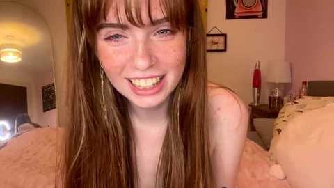 Media: Video of a young Caucasian woman with long, straight brown hair, fair skin, and freckles, smiling broadly. She has a slim build and is indoors, in a cozy bedroom with light walls, a bed, and a red lamp.